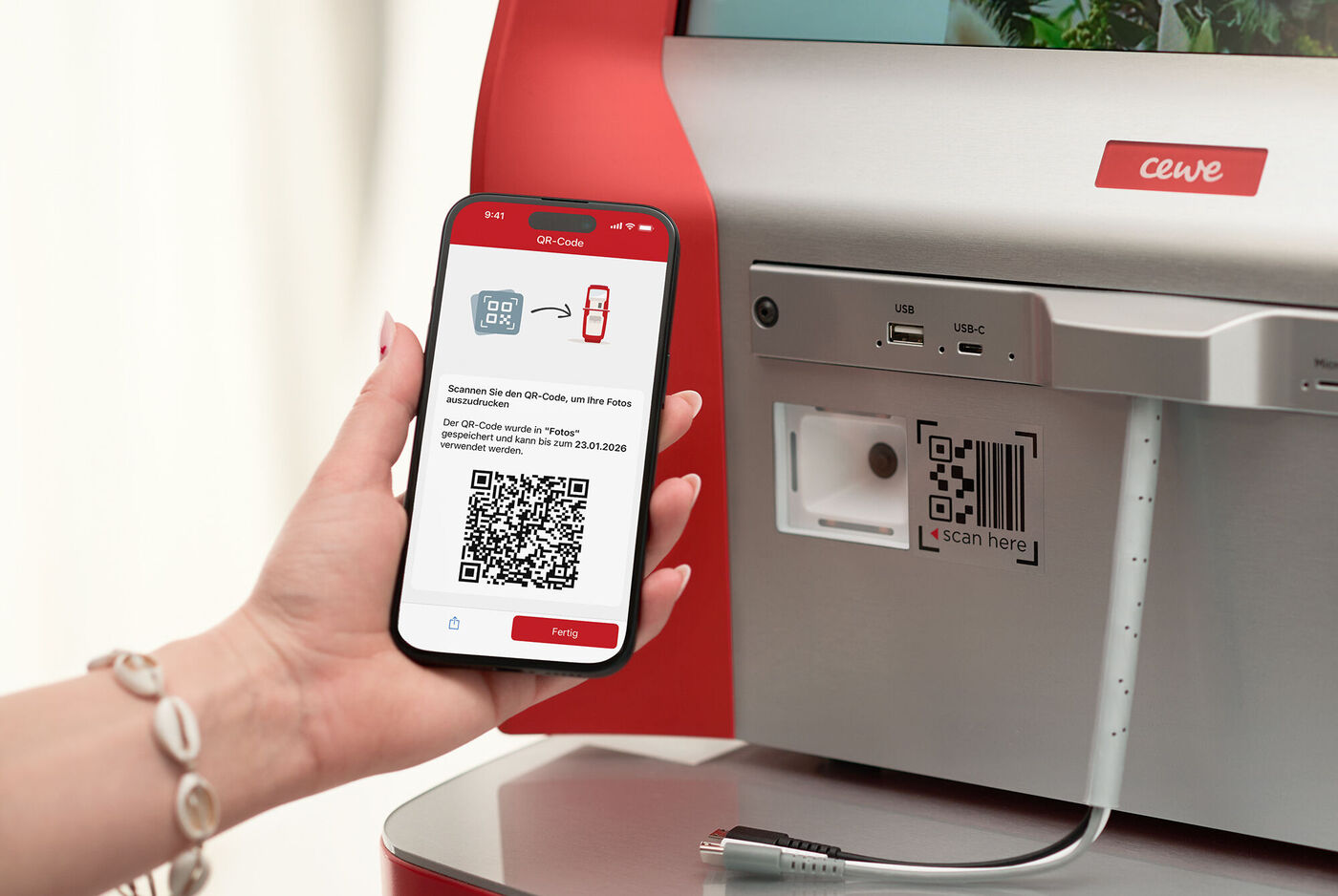 Eine Hand hält ein Smartphone mit einem QR‑Code zum Ausdrucken von Fotos vor einer CEWE Fotostation.